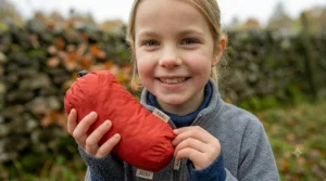A joyful photograph of the girl in a grey fleece, proudly holding a red kids' waterproof jacket that has been compressed into its small, attached stuff sack. It demonstrates the product's innovative packable design, showing how compact and easy it is to carry when not in use.